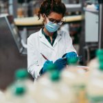 Young happy female worker with protective face mask working in bottling factory and checking water bottles before shipment. Inspection quality control.