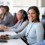 Portrait of smiling businesswoman wearing headset while using computer. Confident female operator is working with colleagues at desk. They are in call center.